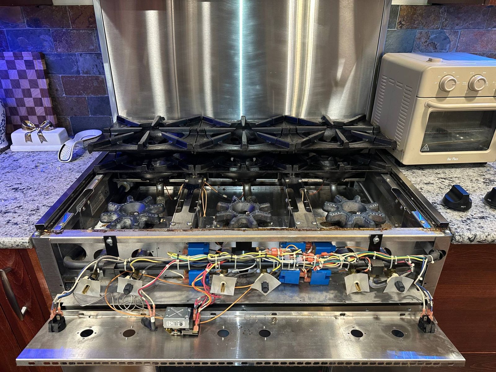 BlueStar range with the front control panel tipped down, showing the burner valves, wiring harness and spark modules behind it