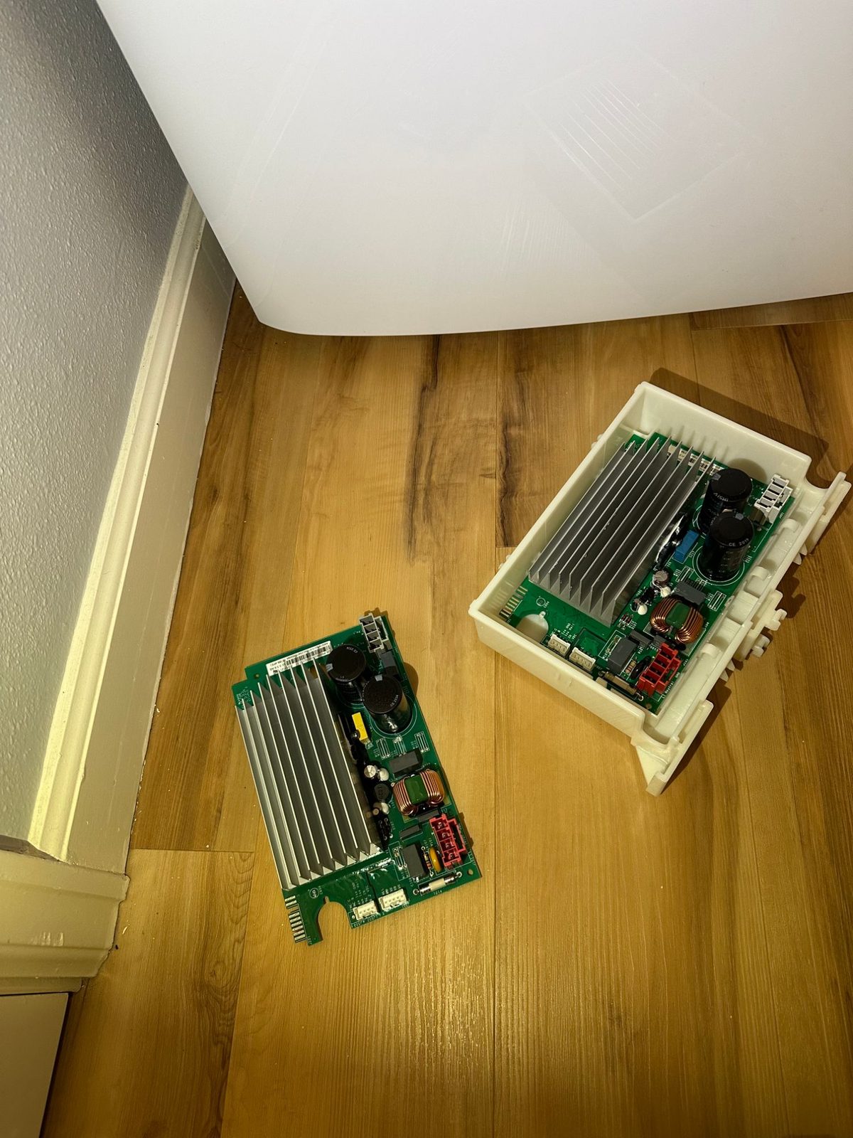 Two GE washer inverter boards on the floor, one loose and one in its plastic housing, both with aluminum heat sinks