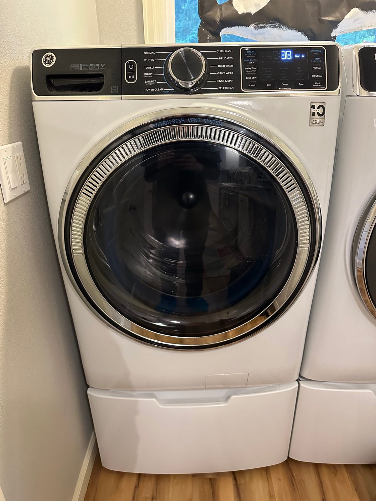 White GE front-load washer on its pedestal in a Lynnwood laundry room, running a cycle after the repair