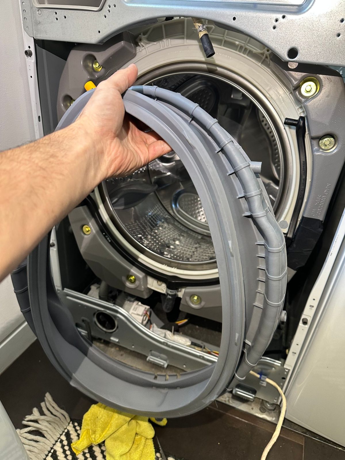 Technician holding a new gray LG door boot gasket with its built-in spray hoses in front of the bare washer tub