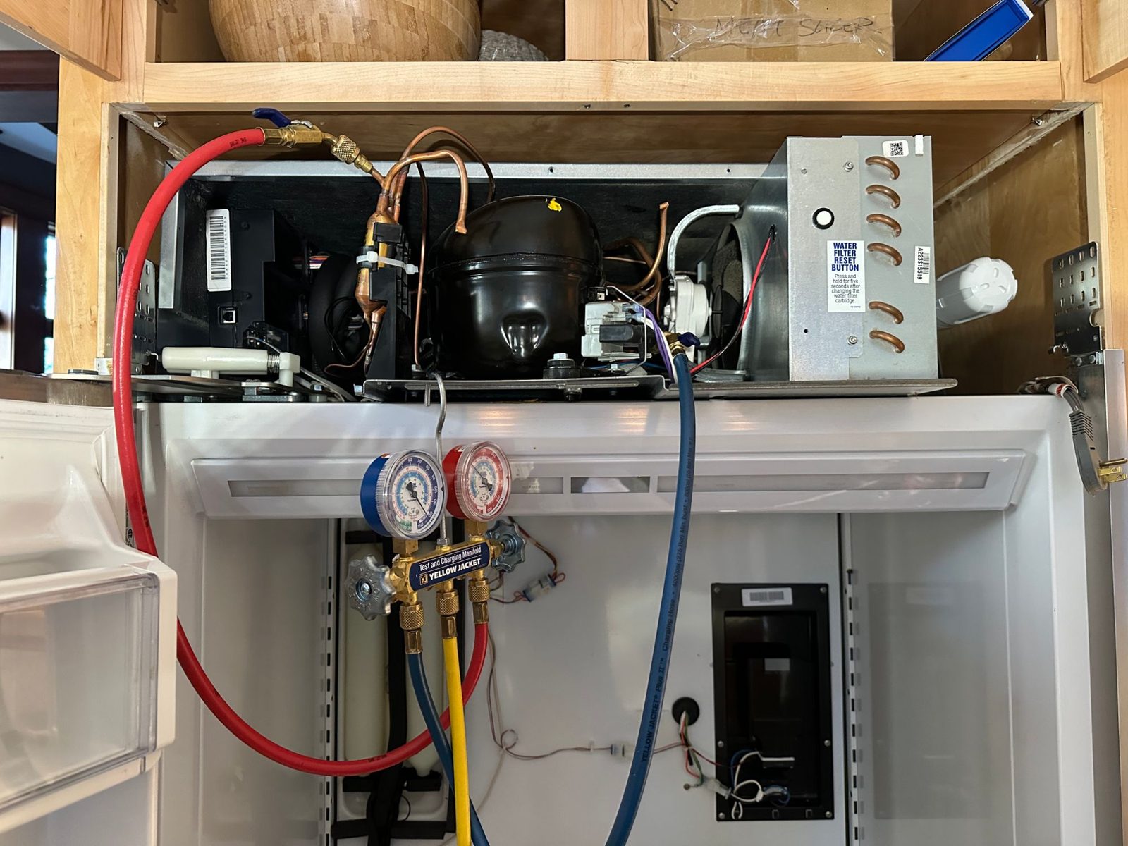Sub-Zero top machine compartment open above the refrigerator, compressor and condenser visible, with refrigeration gauge hoses connected to the sealed system