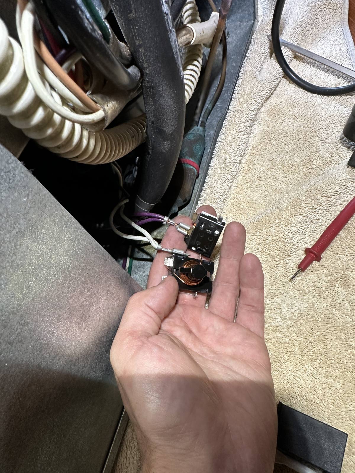 Failed start relay and overload protector removed from the Sub-Zero compressor terminal, held in hand next to a multimeter probe