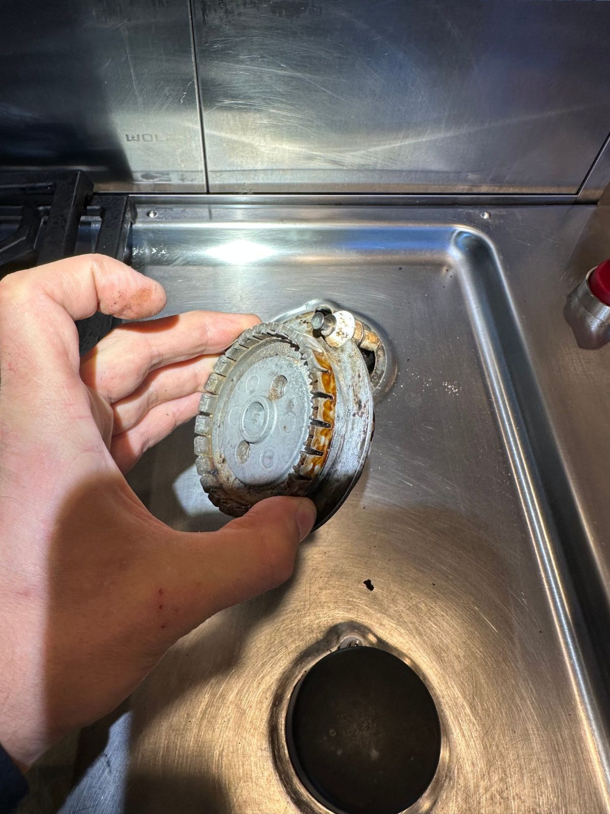 Old Wolf cooktop burner lifted out of the burner pan, showing rust and corrosion around the burner base and igniter