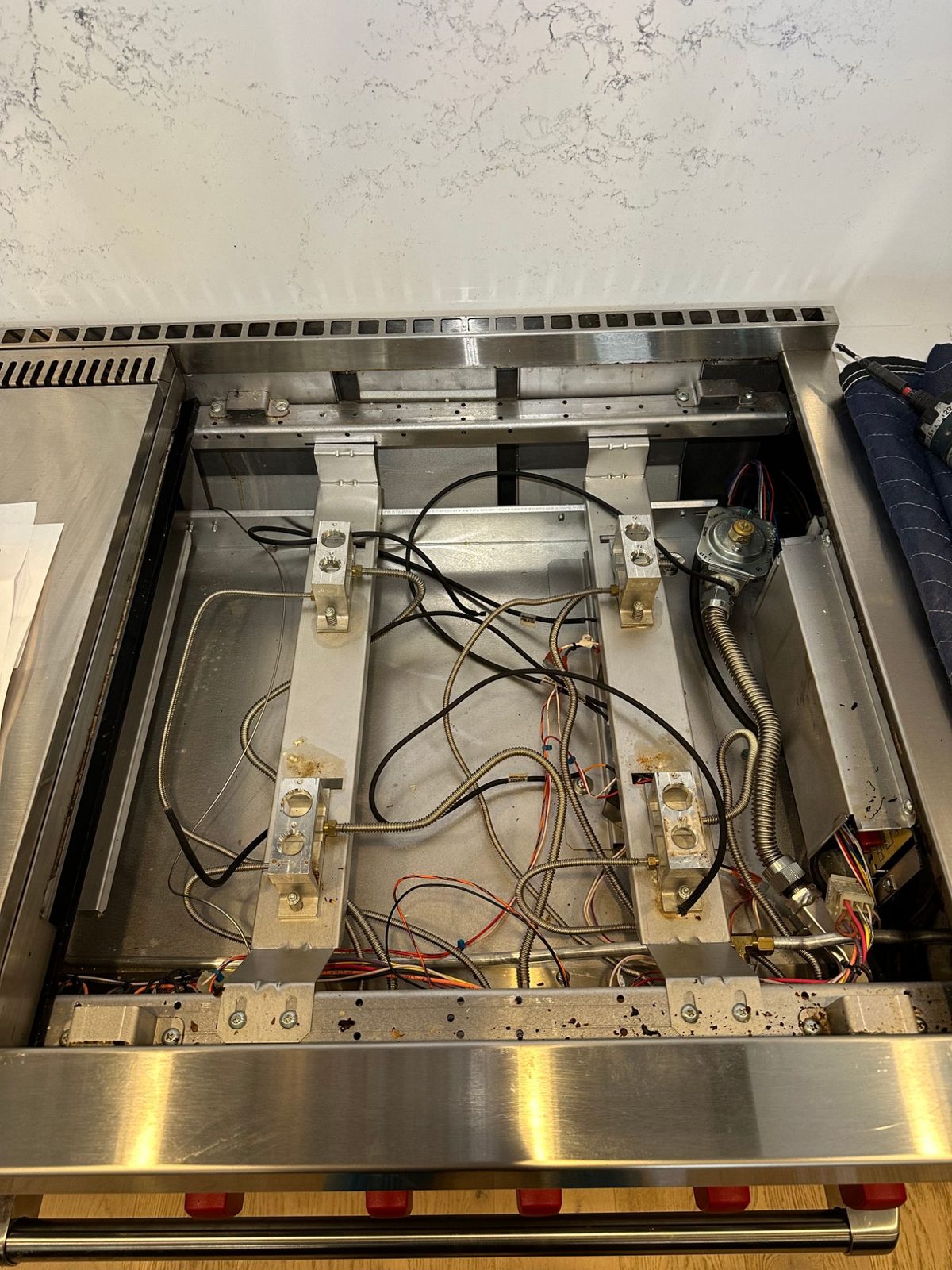 Top-down view inside the Wolf range burner box showing the four burner bases, electrode wires, gas lines, and pressure regulator
